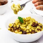 Eine Gabel mit veganen Mac and Cheese (Käsenudeln) über einer weißen Schüssel mit vollwertigen Macaroni und Käse