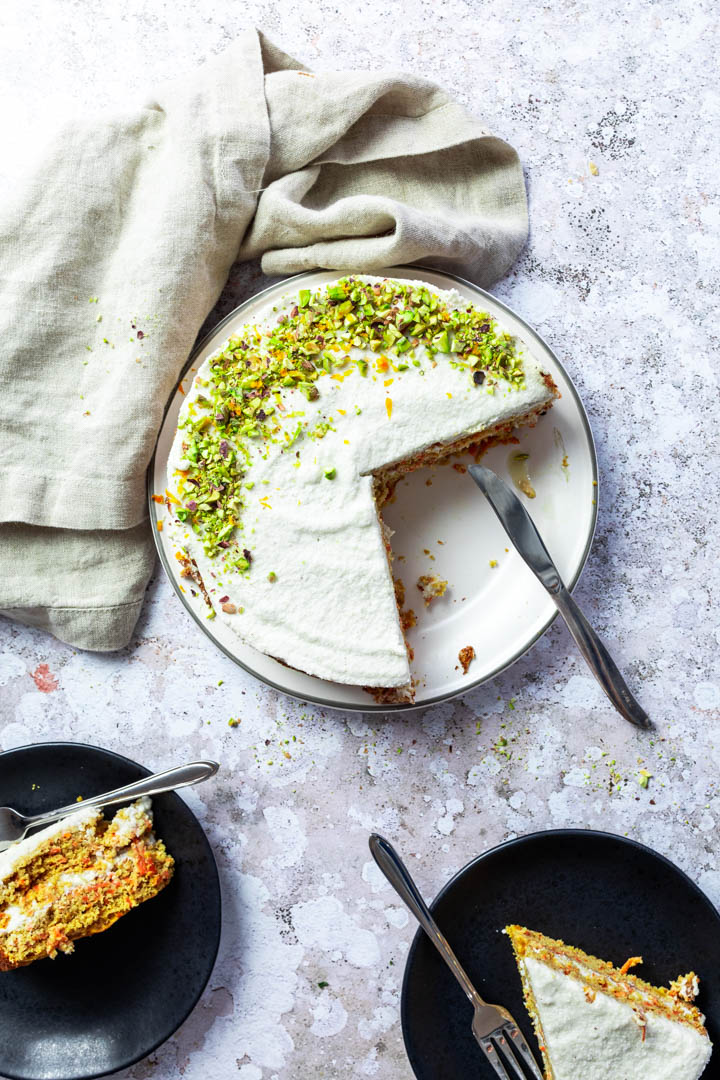 flatlay of a vegan carrot cake decorated with chopped pistachios. Two pieces cut and layed on a plate.