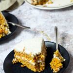 One Piece of Vegan Carrot Cake on a plate with a fork. The fork has some of the carrot cake on top. In the back the whole carrot cake.