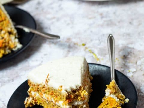 One Piece of Vegan Carrot Cake on a plate with a fork. The fork has some of the carrot cake on top. In the back the whole carrot cake.