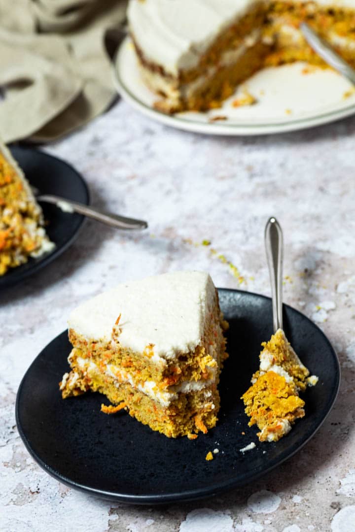 One Piece of Vegan Carrot Cake on a plate with a fork. The fork has some of the carrot cake on top. In the back the whole carrot cake.