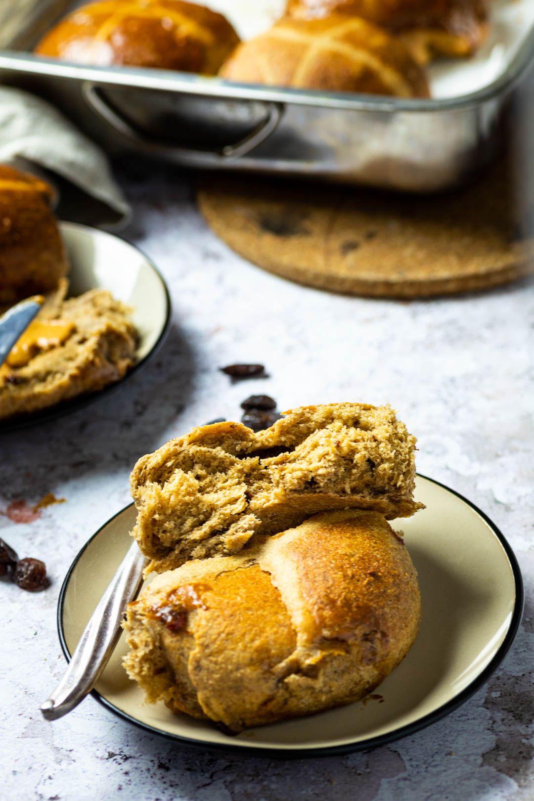 One halfed vegan hot cross bun on a plate with the bakind dish full with hot cross buns in the background