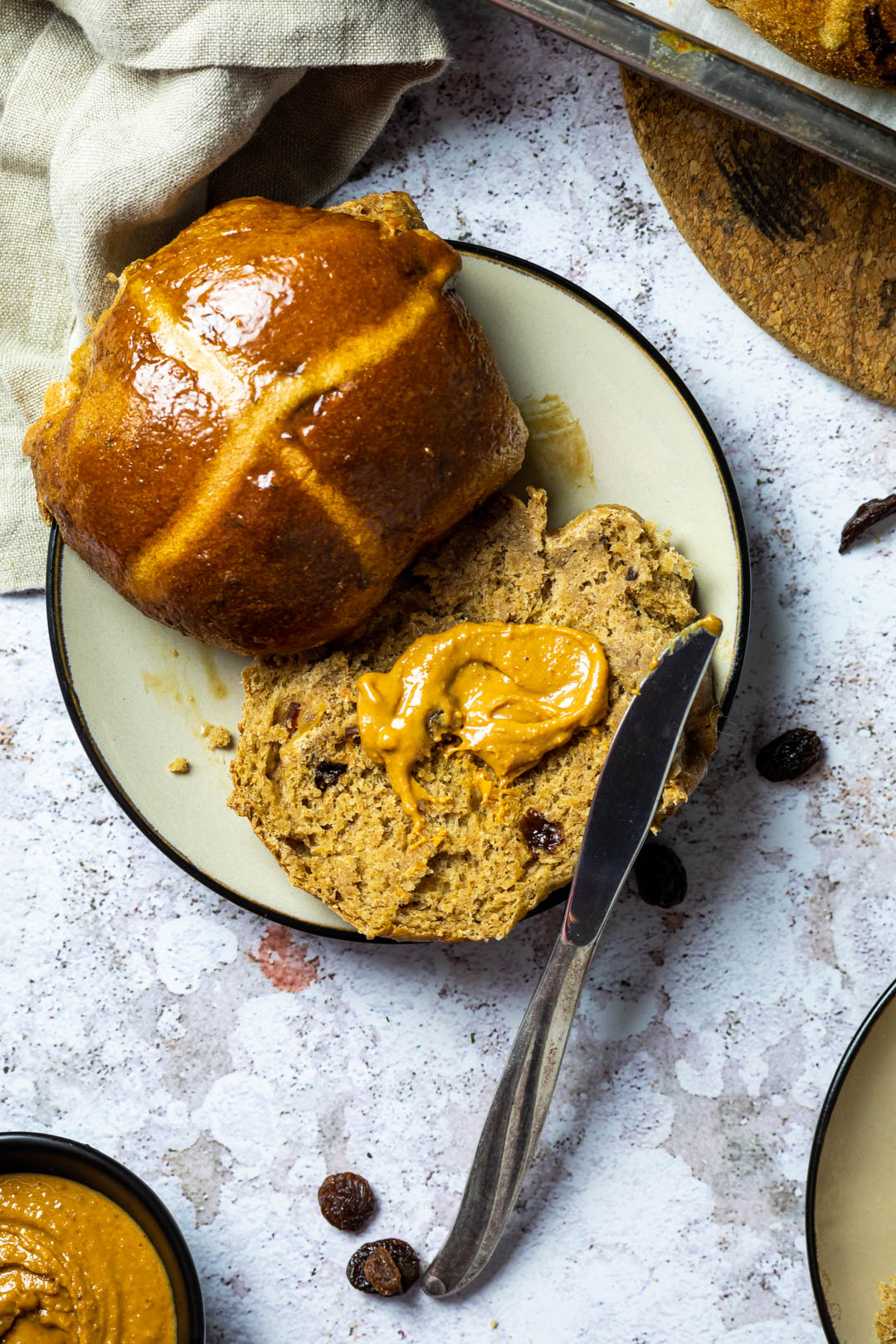 Close up of one vegan hot cross bun which is cut in half and one half is spread with peanut butter