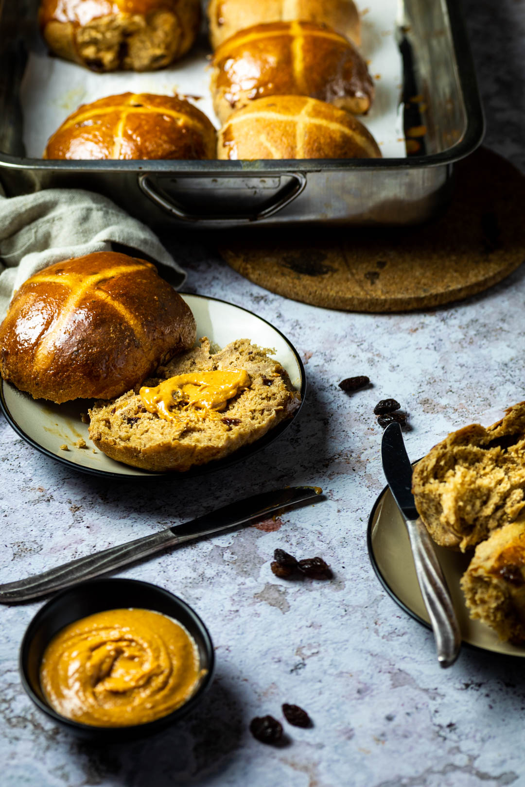 Ein Teller mit einem halbierten veganen Hot Cross Bun in der Mitte des Bildes. Im Hintergrund die Backform mit Hot Cross buns und davor eine kleine Schlüssel mit Erdnussbutter