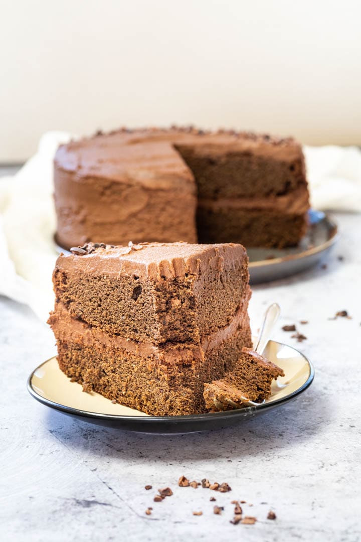 Vegane Schokotorte mit einer Gabel auf einem Teller. Im Hintergrund die ganze vegane Schokoladentorte