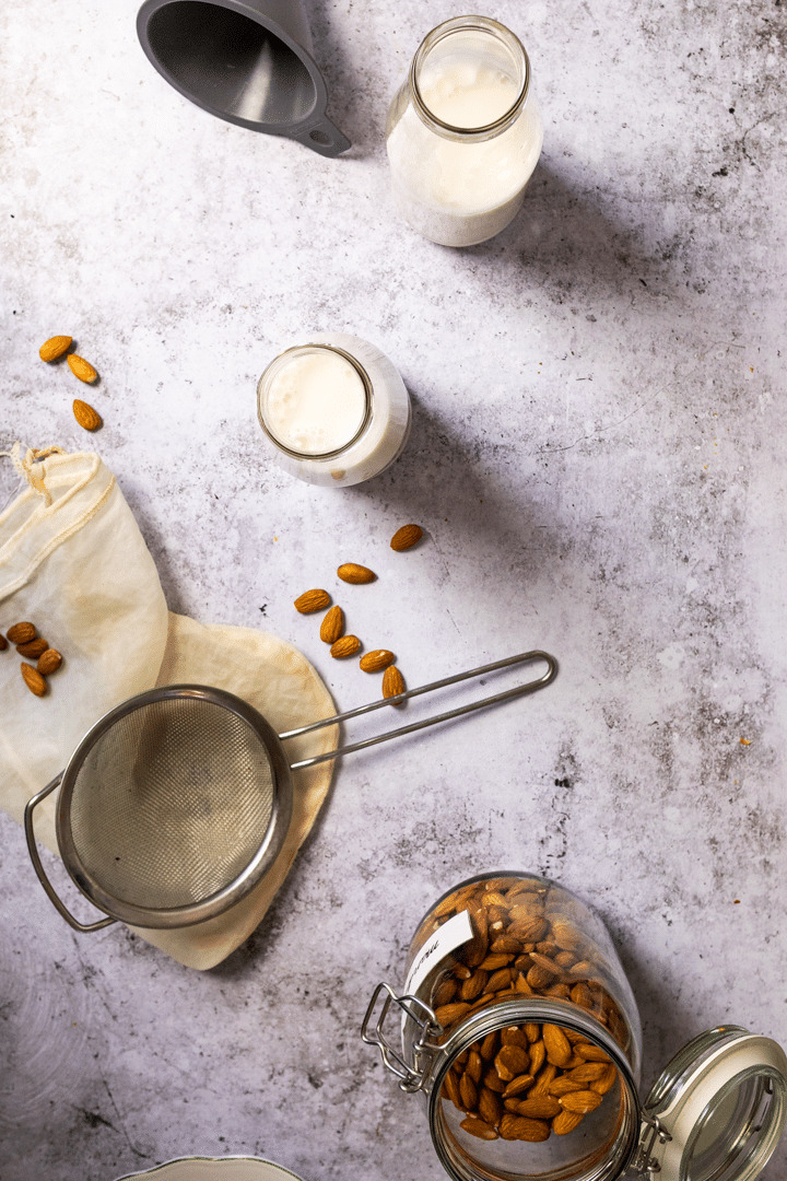 Set up of making homemade almond milk with a jar of almonds, sieve, nut milk bag, and two jars with almond milk