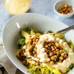 A bowl full with vegan caesar salad. Lettuce topped with crispy chickpeas, vegan parmesan and some caesar dressing.