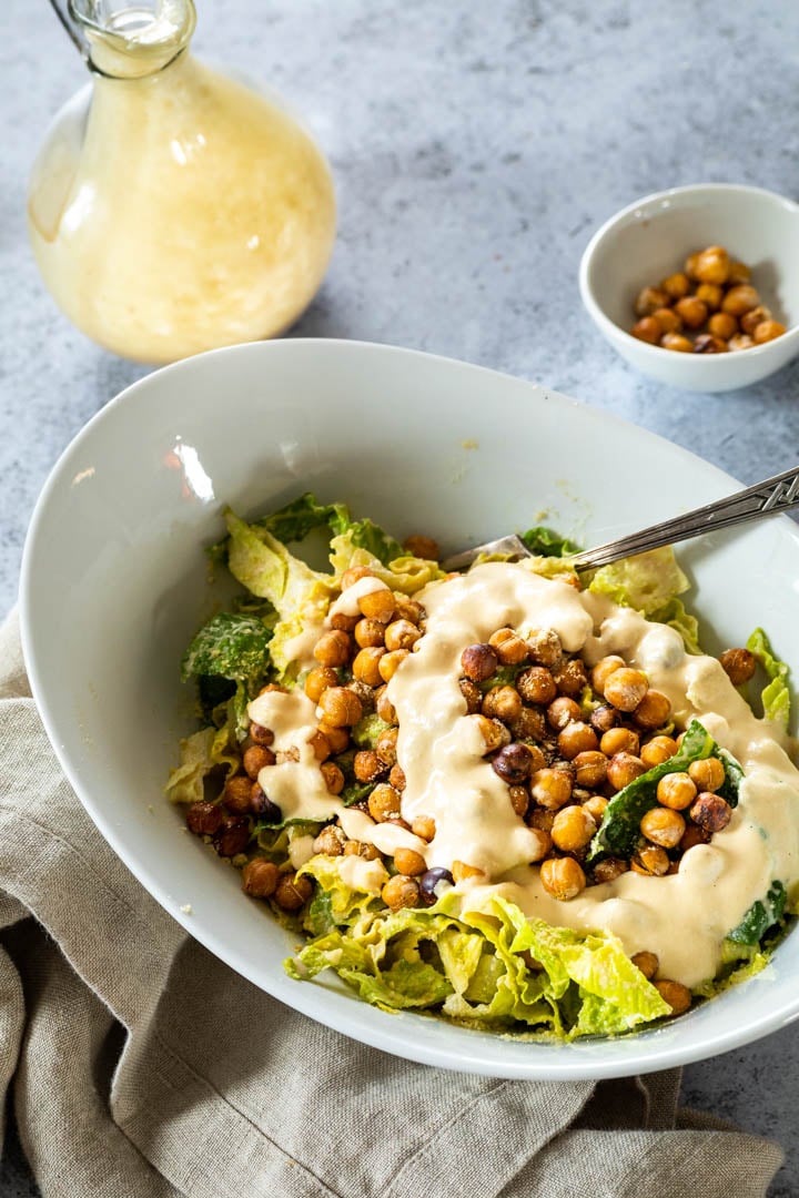 A bowl full with vegan caesar salad. Lettuce topped with crispy chickpeas, vegan parmesan and some caesar dressing.