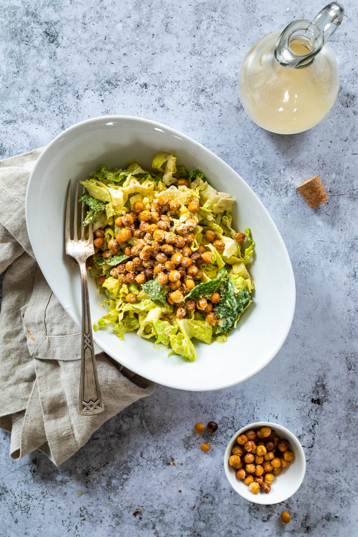 A bowl with a vegan caesar salad in the middle. Above a bottle with the caesar dressing and below a little bowl with crispy chickpeas