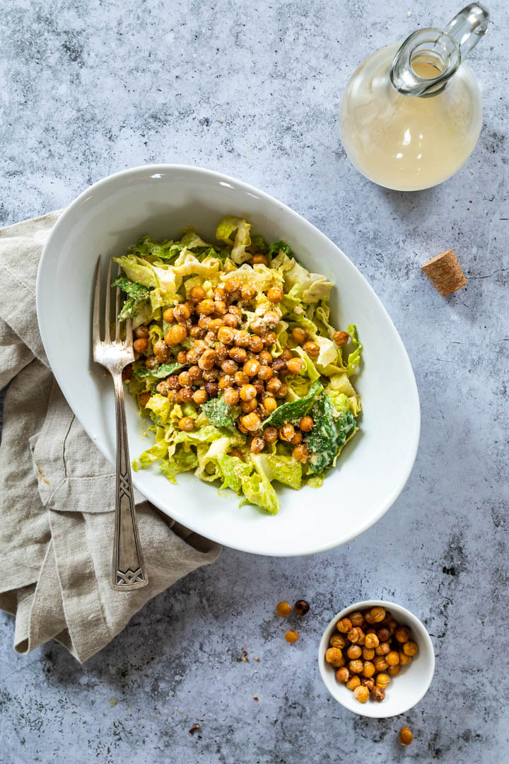 Eine Schüssel mit veganem Caesar Salat in der Mitte und eine Flasche mit Caesar Dressing darüber und darunter eine Schälchen mit knusprigen Kichererbsen.