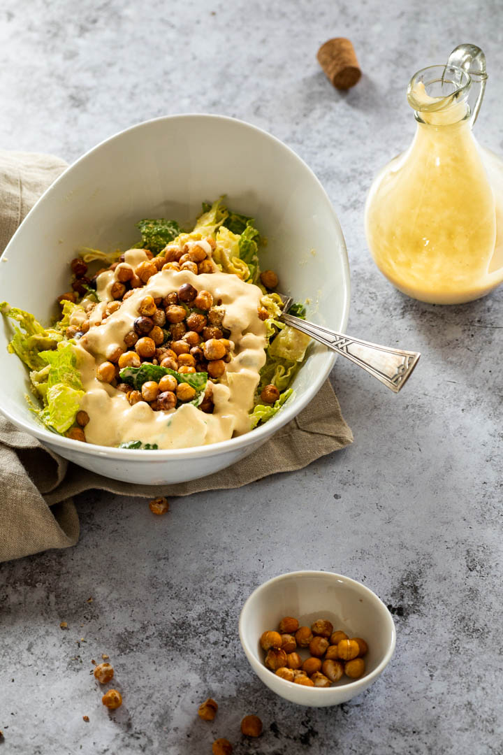 Caesar Salad topped with caesar dressing and a dressing bottle next to the bowl . Below a little bowl with crispy chickpeas