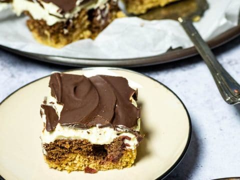 A piece of the vegan custard cherry chocolate cake (German Donauwelle) on a plate with a platter of the cake in the background and one piece in front.