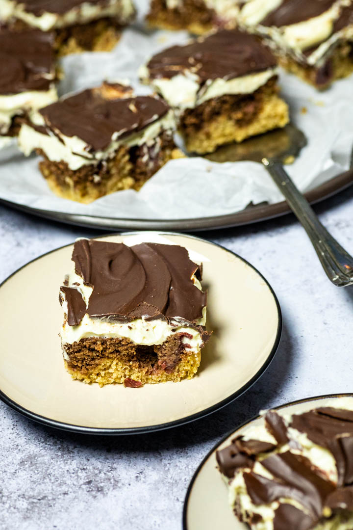 A piece of the vegan custard cherry chocolate cake (German Donauwelle) on a plate with a platter of the cake in the background and one piece in front.