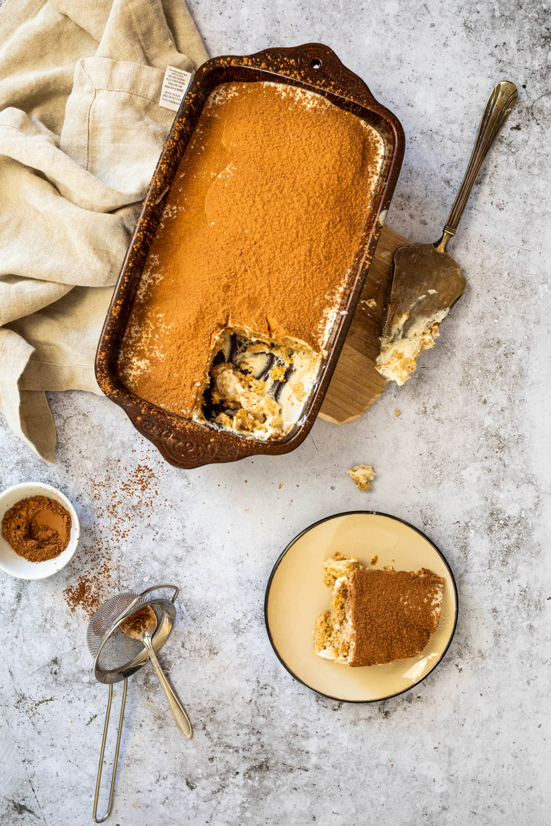 Veganes Tiramisu in einer Pfanne mit einem Stück auf einem Teller und Kakaopulver in einer kleinen Schüssel.