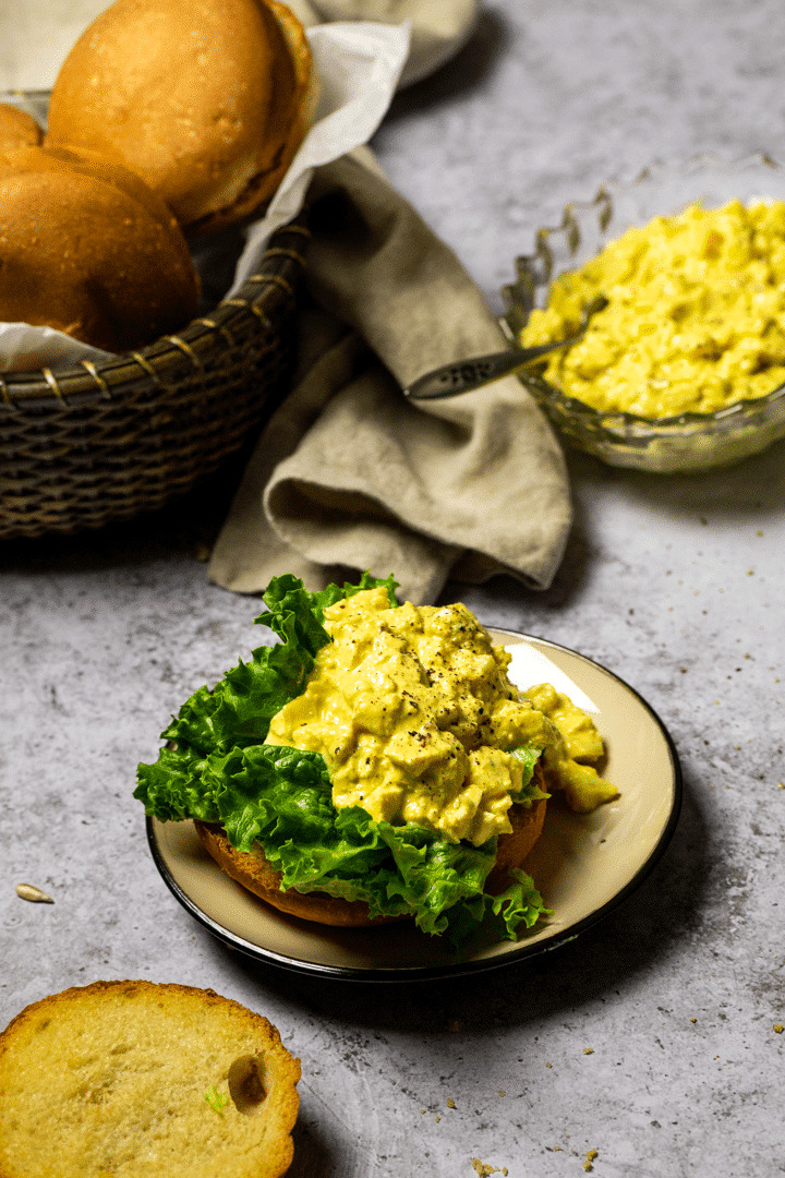 Vegan Egg Salad on a bun in the background buns in a basket and egg salad in a bowl