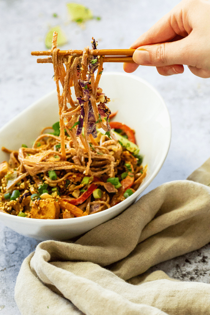 Bowl with some whole food plant-based thai peanut noodles and some are holding on asian sticks