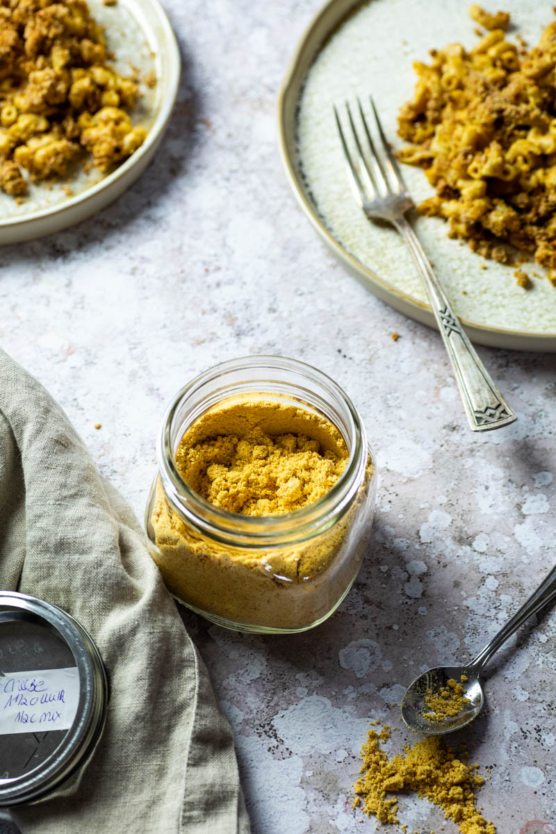 In einem Glas etwas veganes Mac- und Käsepulver mit zwei Tellern mit veganem Mac und Käse im Rücken