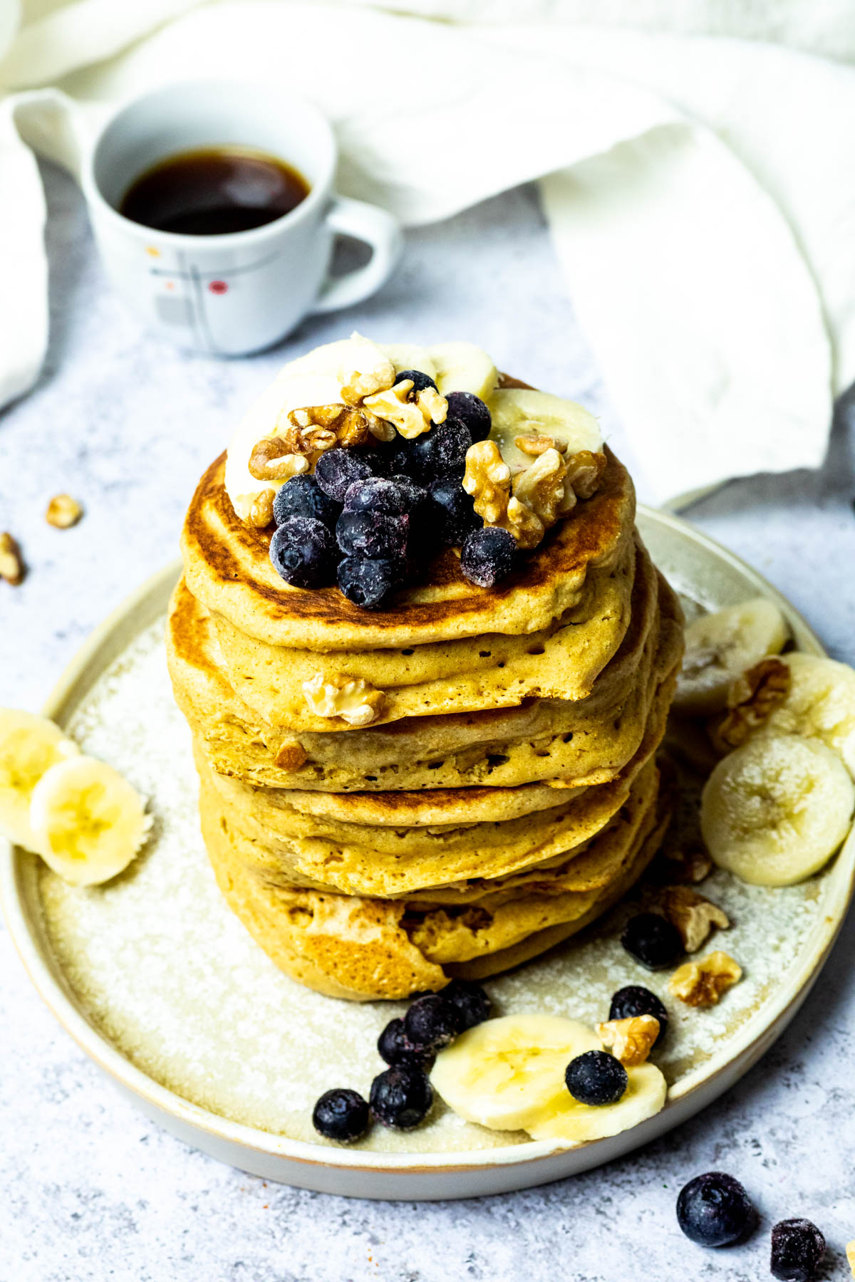 Stack of vegan pancakes on a plate with blueberries, bananas and walnuts. And a cup of coffee in the background