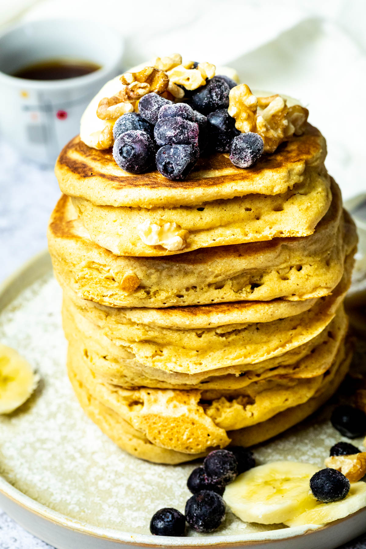 Close up of a Stack of vegan pancakes on a plate with blueberries, bananas and walnuts. And a cup of coffee in the background