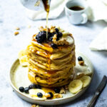 Stack of vegan pancakes on a plate with blueberries, bananas and walnuts. And a cup of coffee in the background. Pouring Maple syrup over the pancake stack.