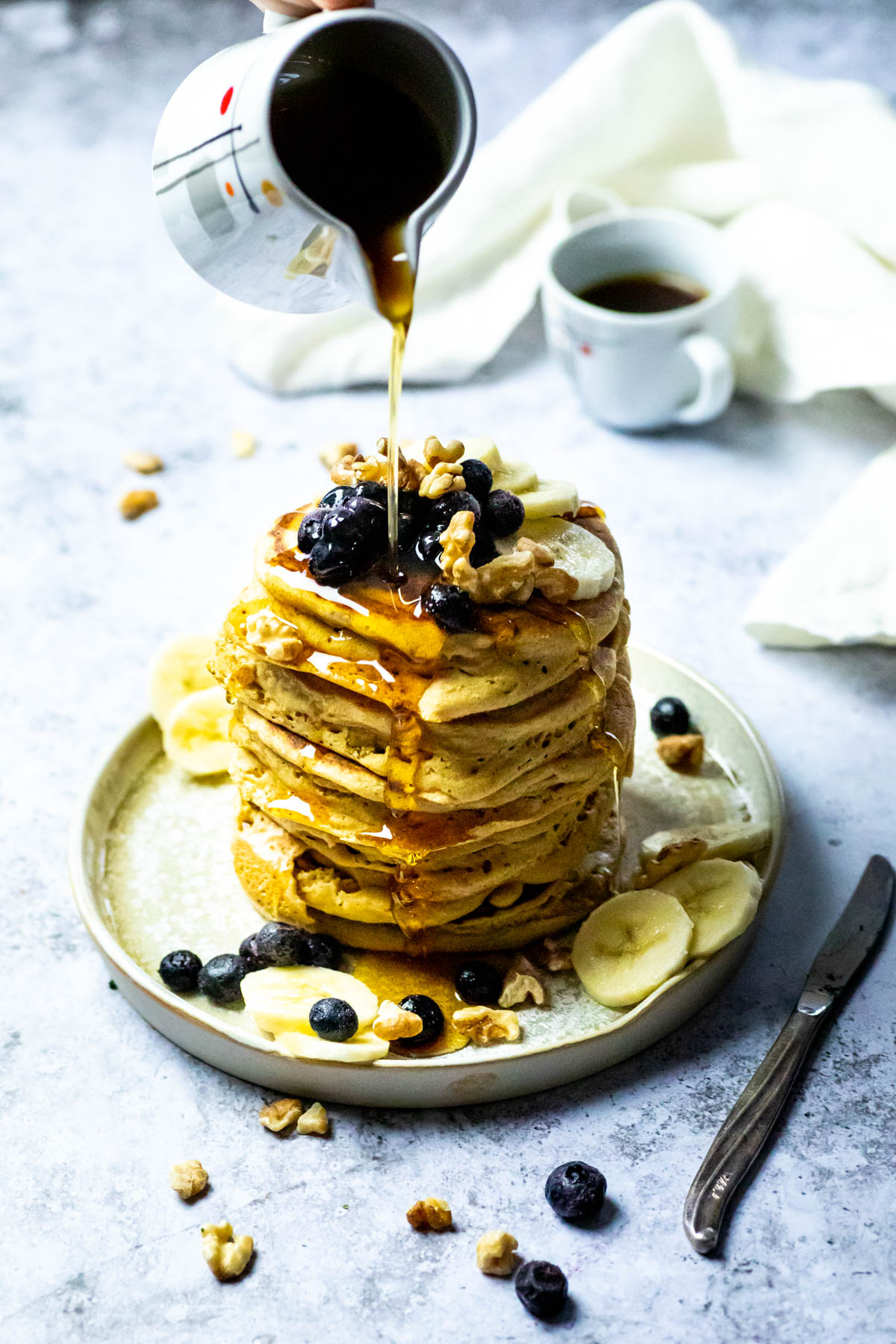 Stack of vegan pancakes on a plate with blueberries, bananas and walnuts. And a cup of coffee in the background. Pouring Maple syrup over the pancake stack.