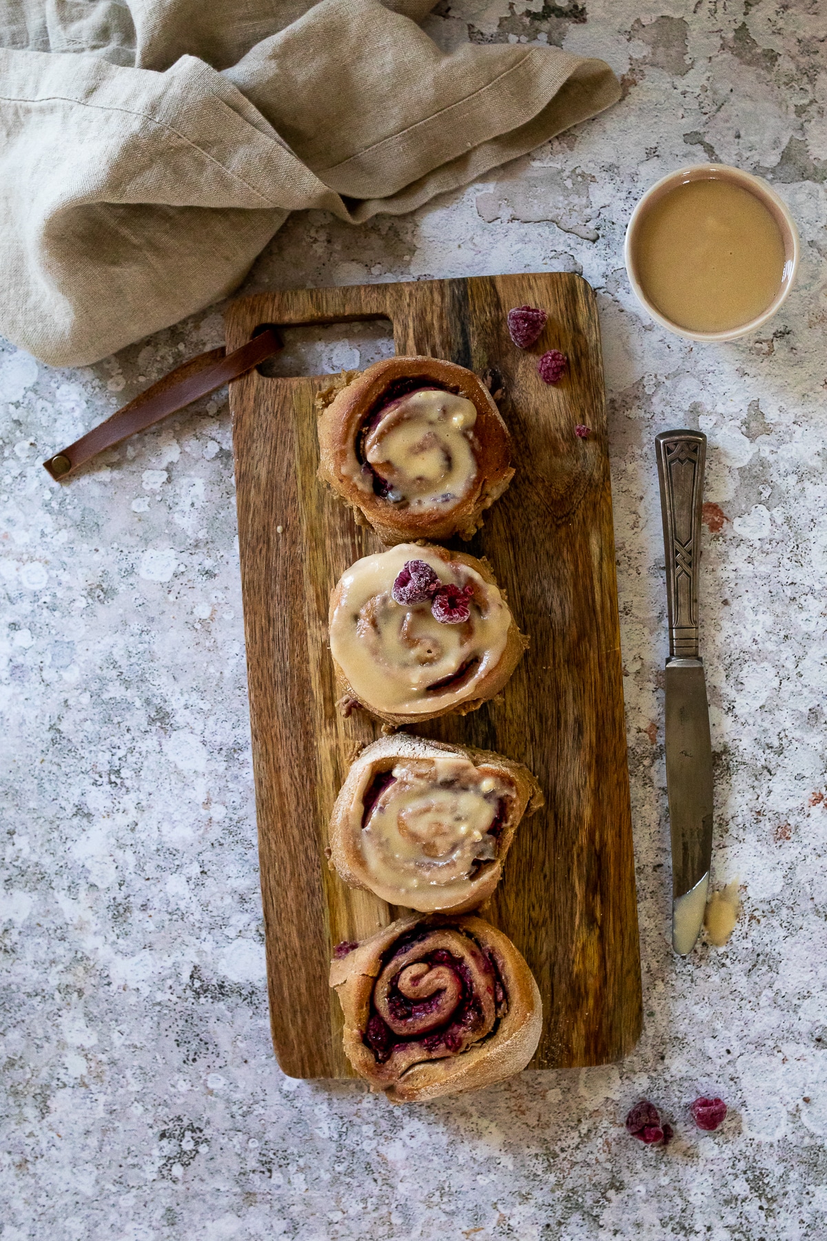 Bird View: 4 vegan raspberry cinnamon buns with an oil free frosting on a wood board with a knive on the right side and a bowl with frosting in the right upper corner.