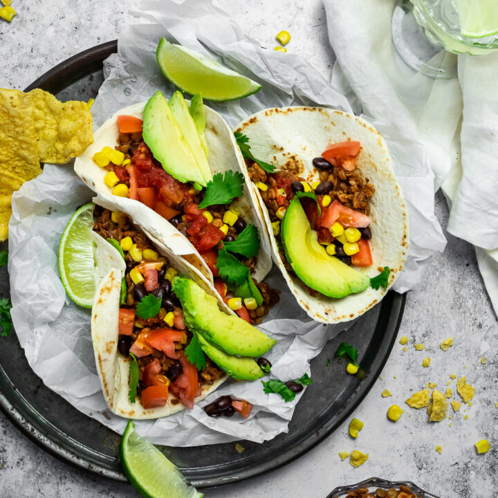 Bird view of 3 tacos on a serving plate with a bowl of vegan taco meat and a cup with water.