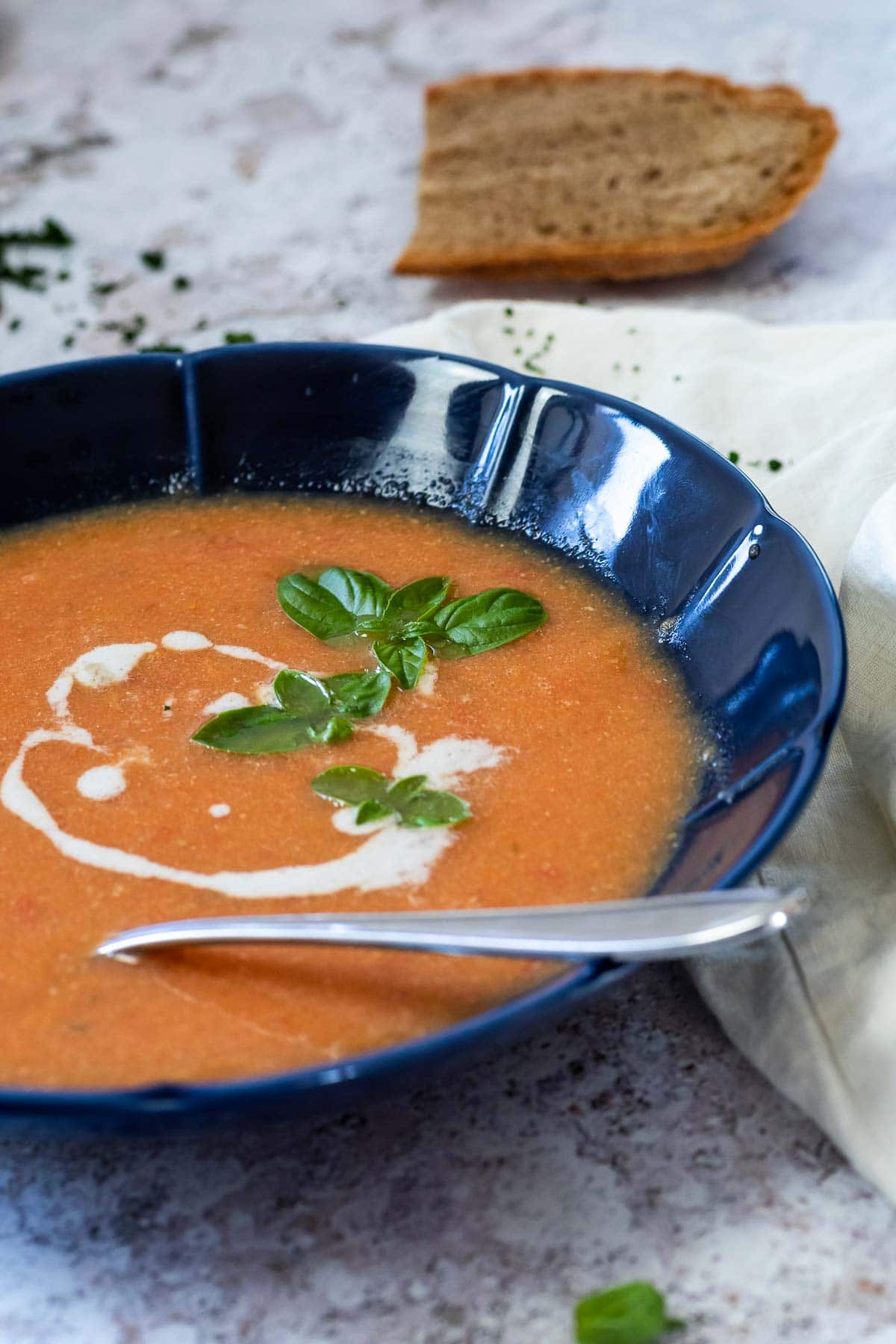 Vegane Tomatensuppe serviert in einer blauen Schüssel mit einem Löffel in der Schüssel. Das Brot liegt verschwommen im Rücken.