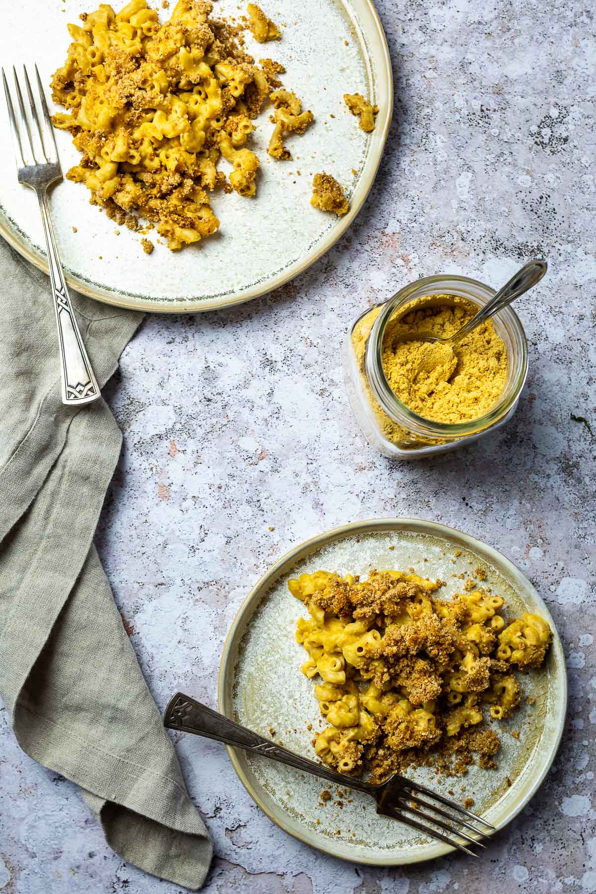 Two plates with baked mac and cheese and in the middle a jar with dairy free baked mac and cheese powder.