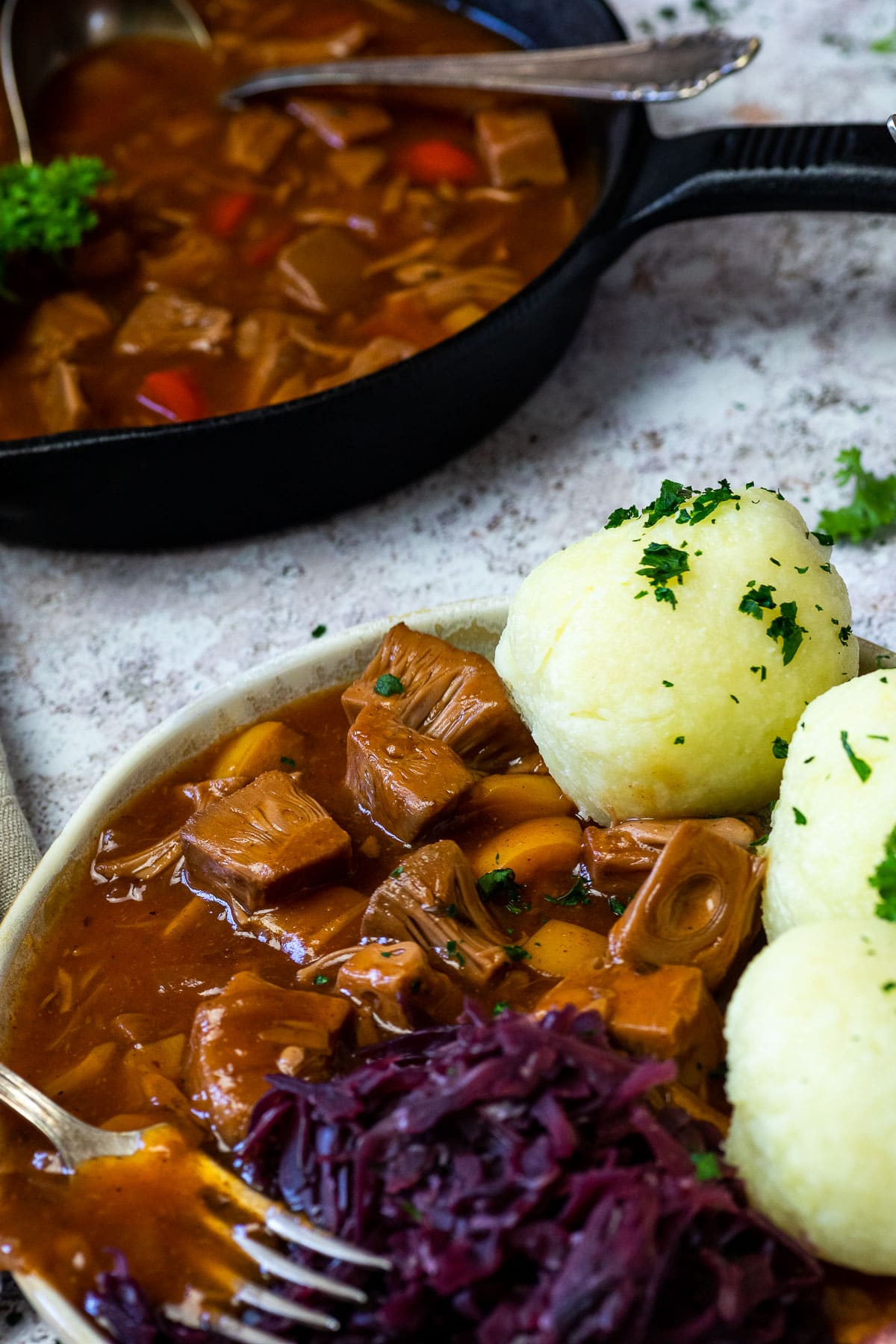 Nahaufnahme des veganen ungarischen Gulasches, das auf einem Teller mit Knödeln und Rotkohl serviert wird. Hinten eine gusseiserne Pfanne.