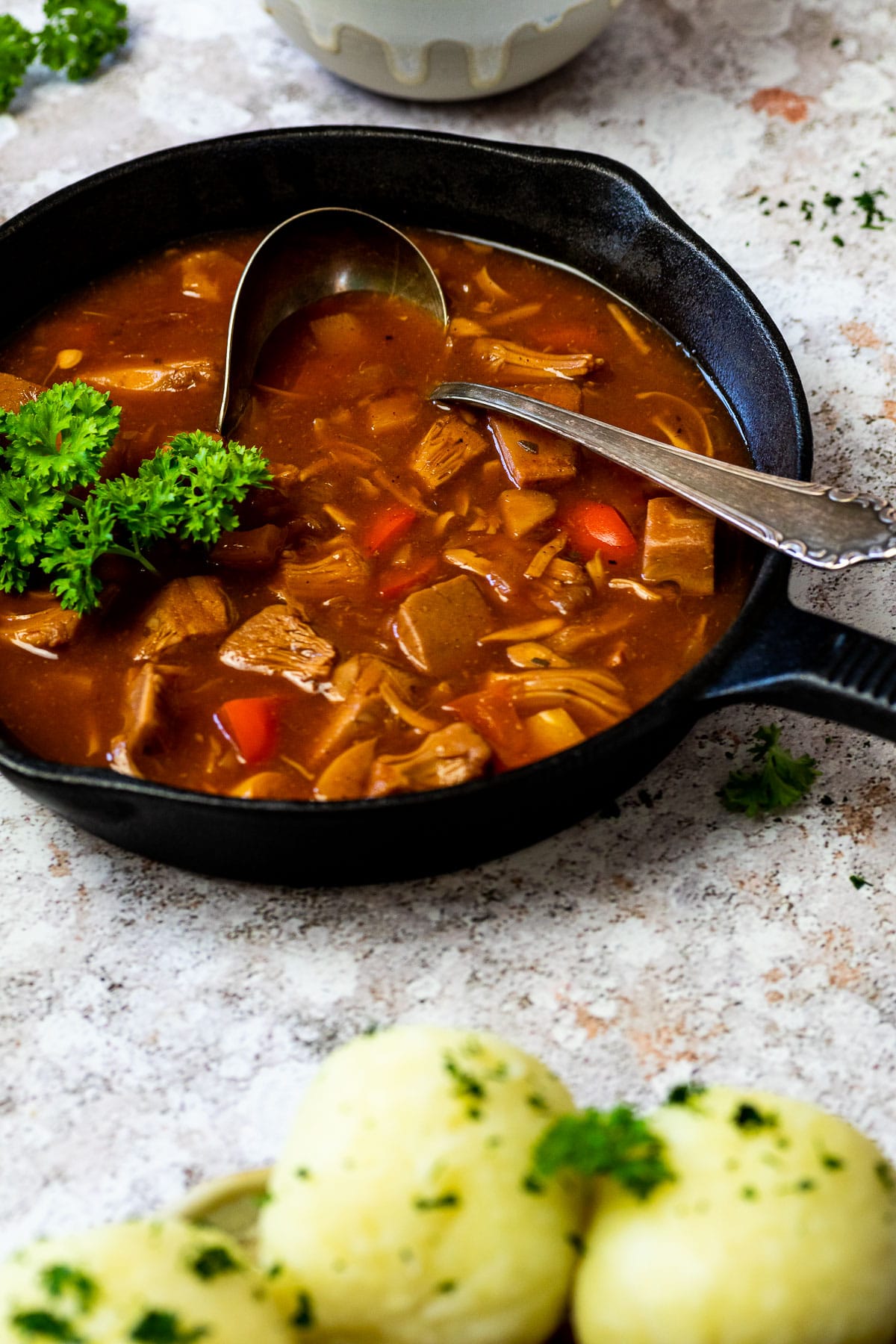 Fleischloses ungarisches Gulasch in einer gusseisernen Pfanne mit verschwommenen Kartoffelknödeln vorne.