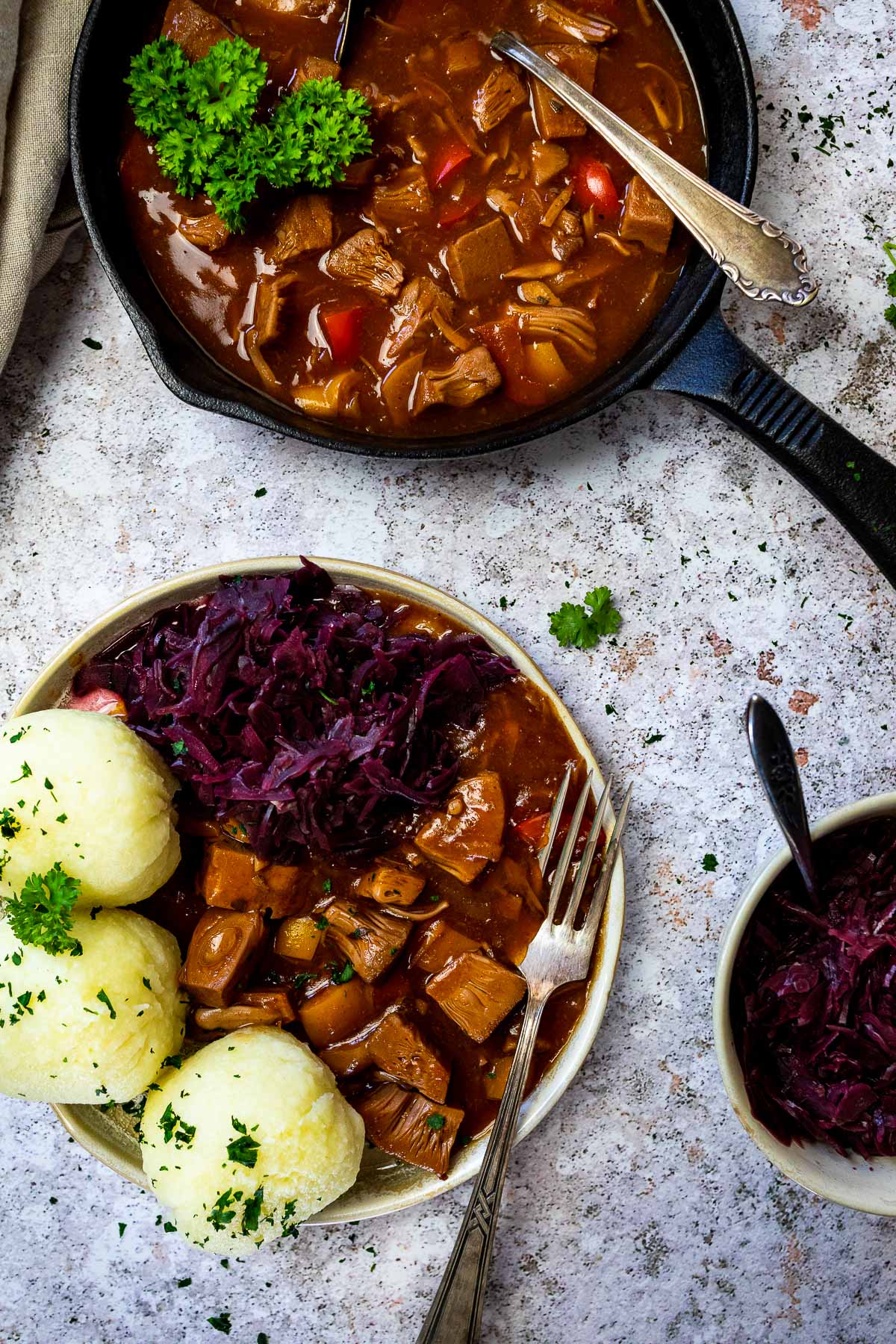 Veganes ungarisches Gulasch serviert mit Kartoffelknödeln und sautiertem Rotkohl und ungarischem Gulasch in einer gusseisernen Pfanne.