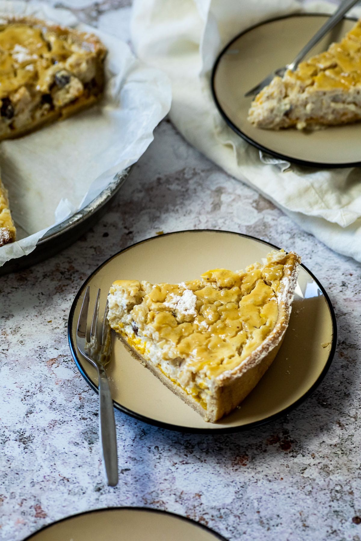 A vegan rice pudding pie piece on a plate with a fork in front of a pie pan and another plate.