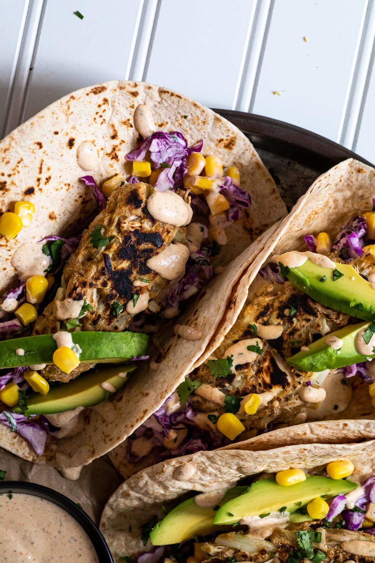 Vegane Fisch-Tacos werden auf einer Platte mit einer Min-Schüssel mit Sauce und einer halben Avocado im Rücken serviert.