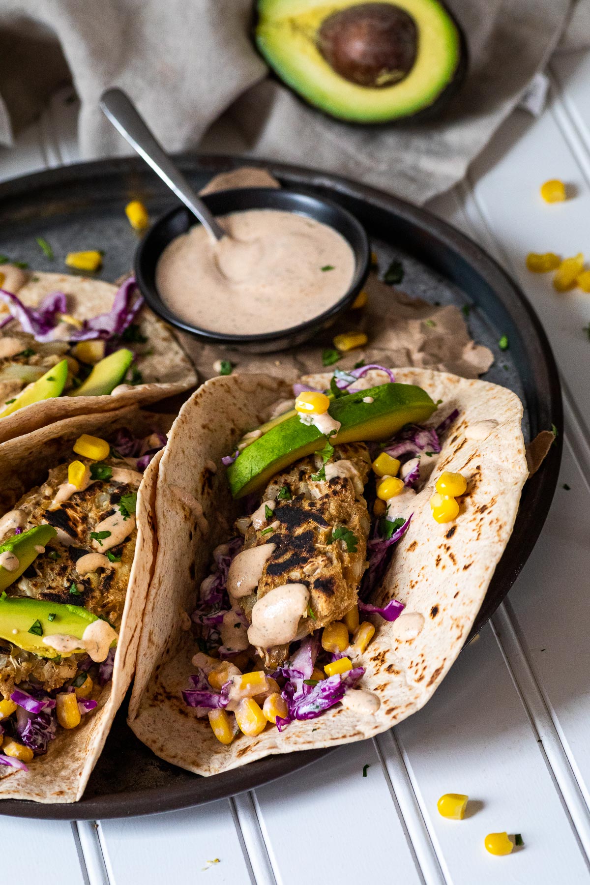 Vegane Fisch-Tacos werden auf einer Platte mit einer Min-Schüssel mit Sauce und einer halben Avocado im Rücken serviert.