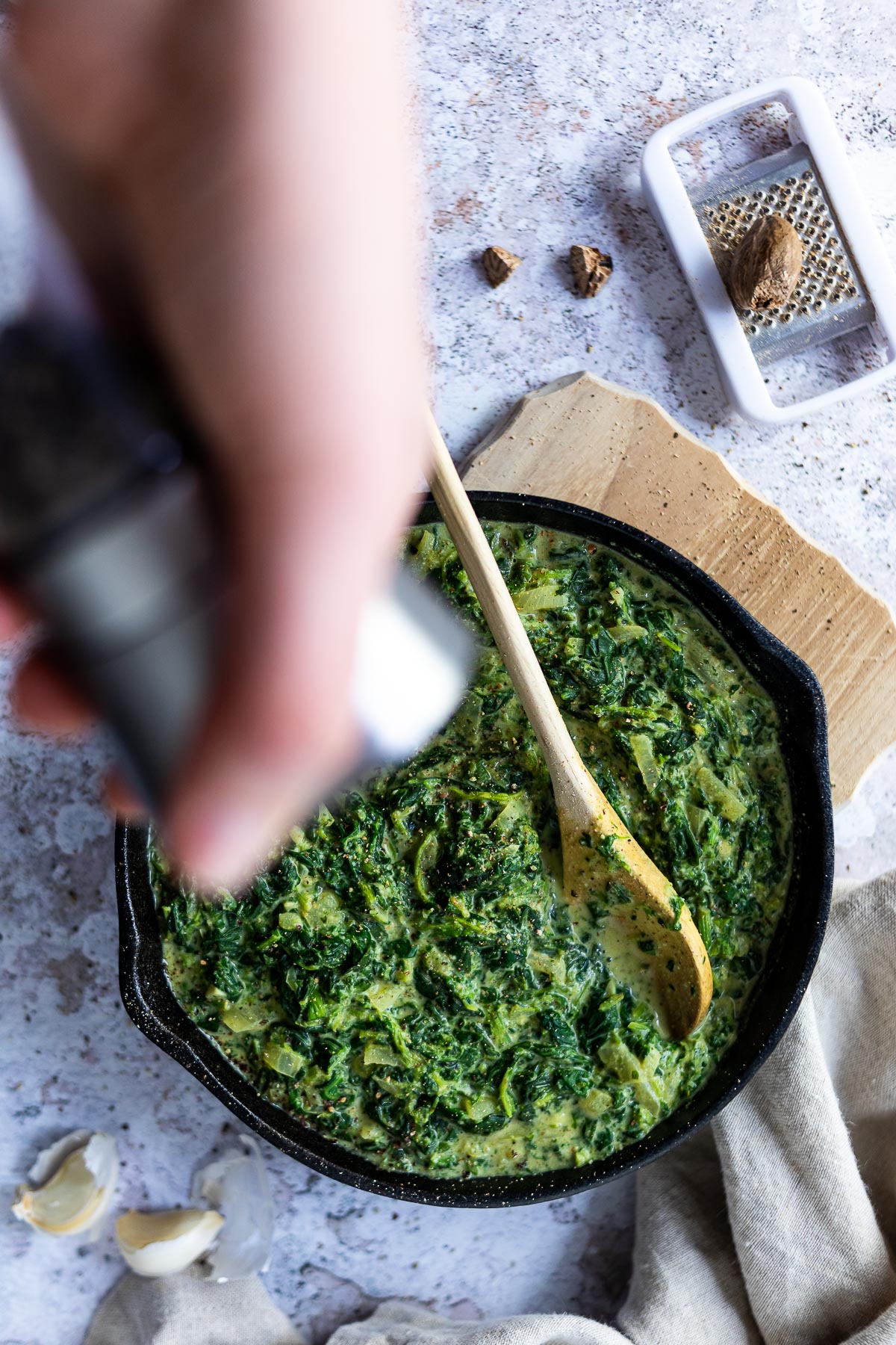 Vegan Creamed Spinach in a cast iron pan with nutmeg on the side. Holding a pepper grinder over the pan blurred.