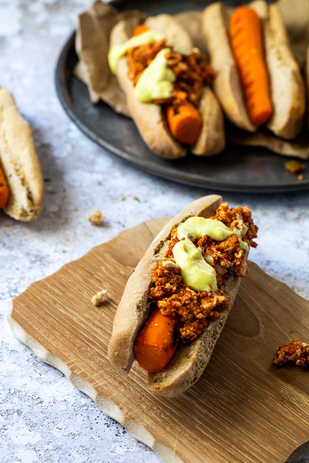 One vegan chili cheese dog on a wooden board and a platter in the background