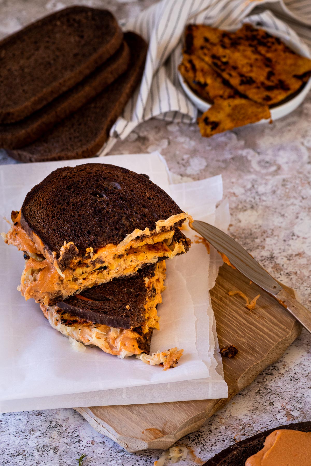 2 vegan Reuben sandwiches on a board with tempeh and bread in the background