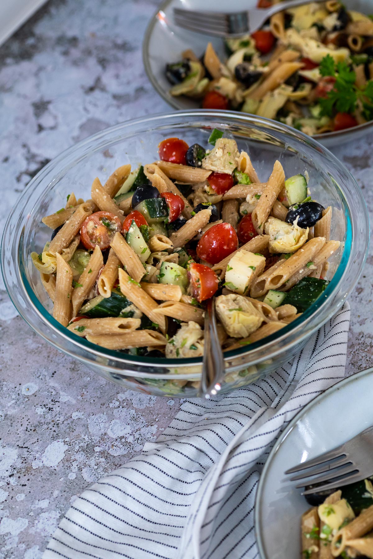 A bowl with vegan artichoke pasta salad and two plates in the background