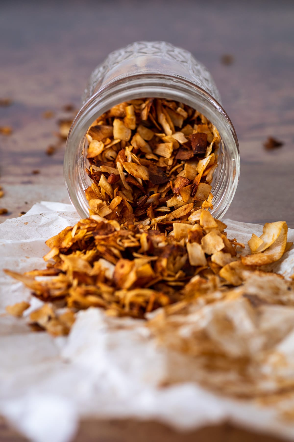 A Jar with coconut bacon is laying on parchment paper with the opening in front. Seeing all the coconut bacon in the jar and some on the paper.