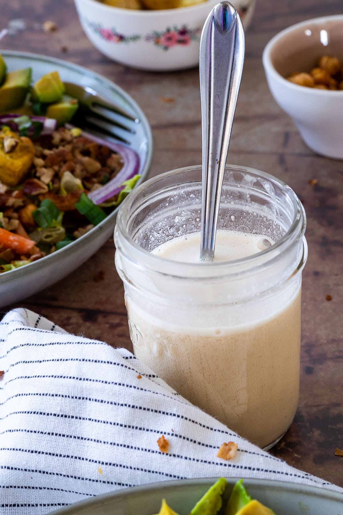 Mason Jar filled with Tahini Dressing and a spoon in the jar. In the background a salad bowl.