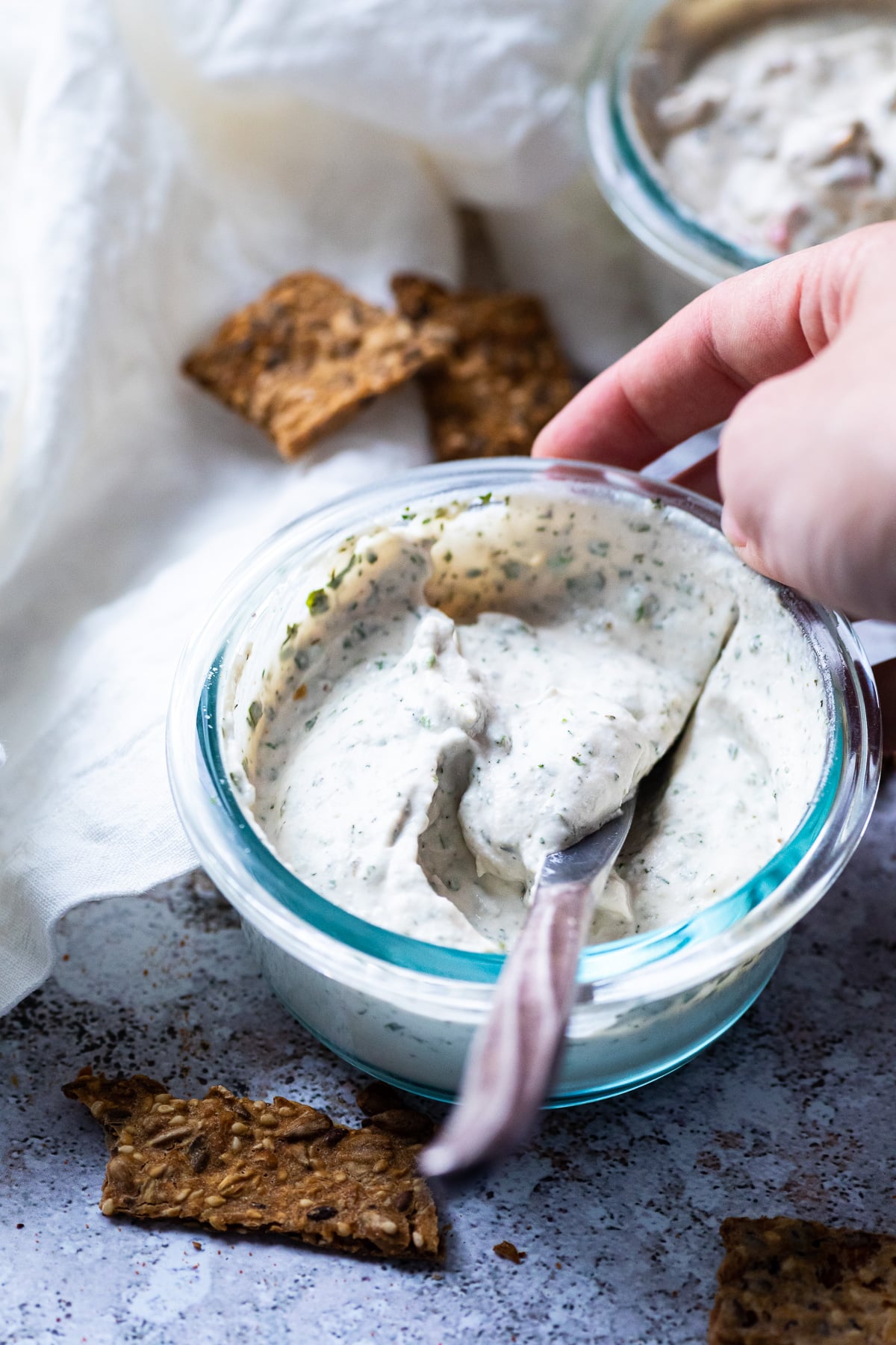 Ein Messer in der Schüssel mit veganem Kräuter-Knoblauch-Frischkäse und Crackern herum.