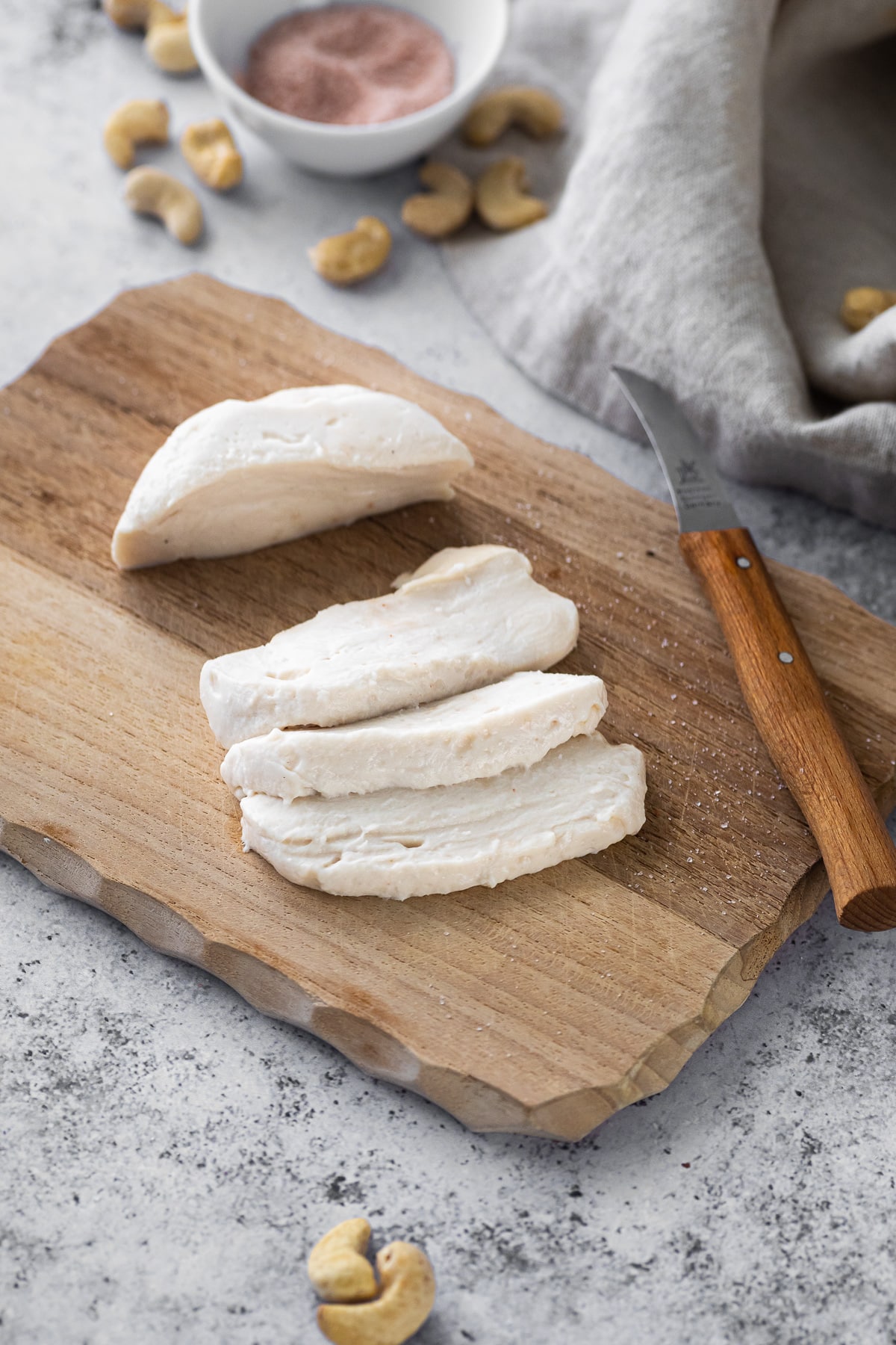 vegan mozzarella cheese sliced on a cutting board with a small knife on the side and some cashews around the board.